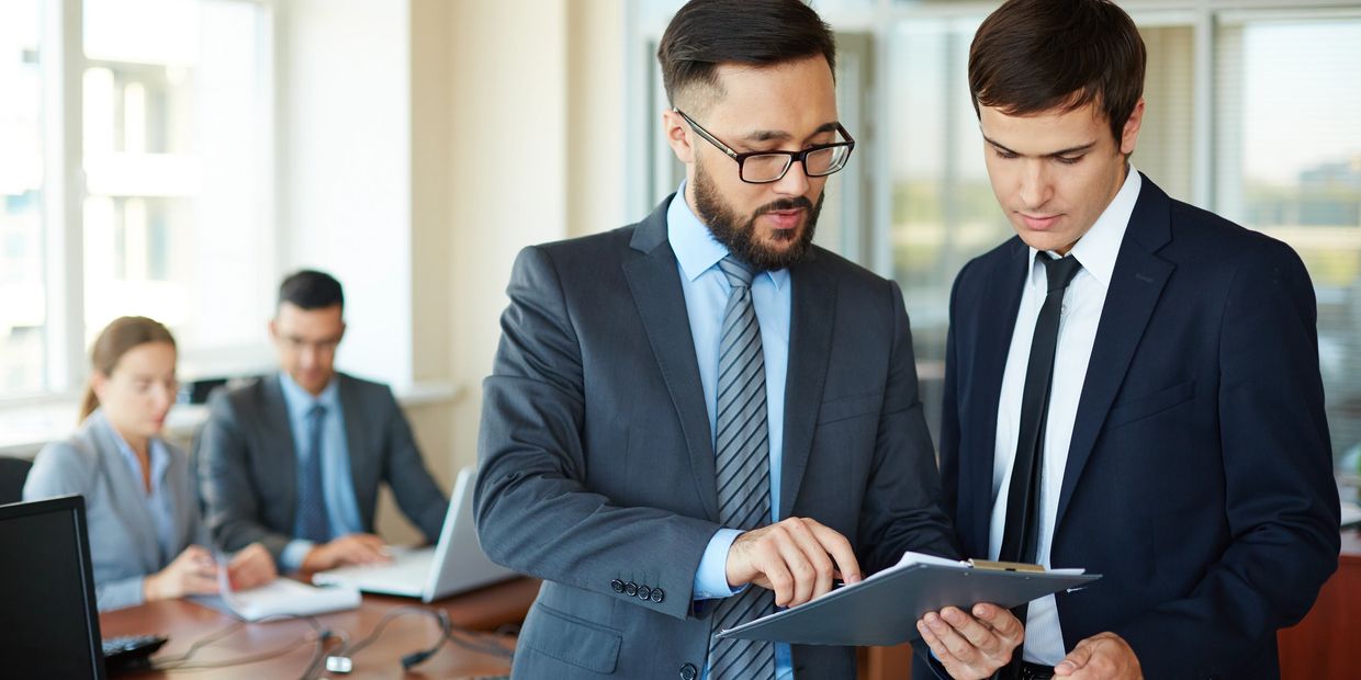 Confident businessman pointing at document while explaining his idea to his partner on background of their colleagues